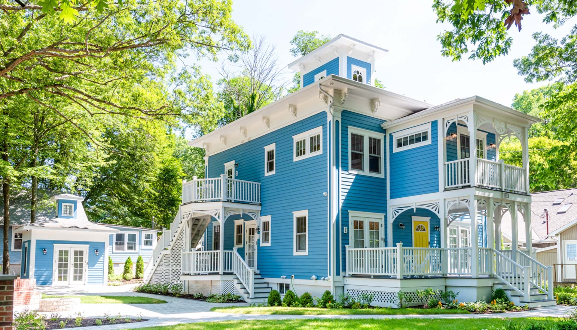 A large, blue multi-story house with white trim and balconies, surrounded by trees and greenery.